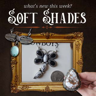 A styled jewelry display featuring multiple silver-toned pieces with black, white, and turquoise stones, arranged inside an ornate gold frame on a dark brown background. A hand holds a large teardrop-shaped stone pendant at the lower right.