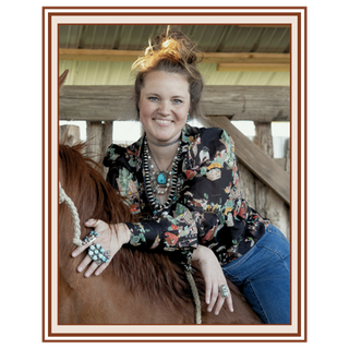 A smiling woman with her hair in a messy bun sits on a brown horse inside a rustic wooden stable. She is wearing a black long-sleeve blouse with a colorful floral and western print, layered with multiple turquoise and silver necklaces, and blue jeans | Texanna Belles Jewelry 